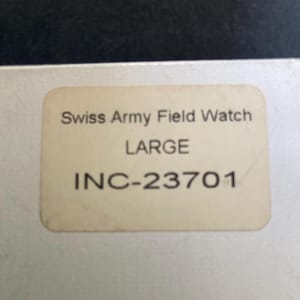 May include: A close-up of a beige label with the text "Swiss Army Field Watch LARGE INC-23701" in black. The label is slightly worn and has rounded corners. The background is a light color.