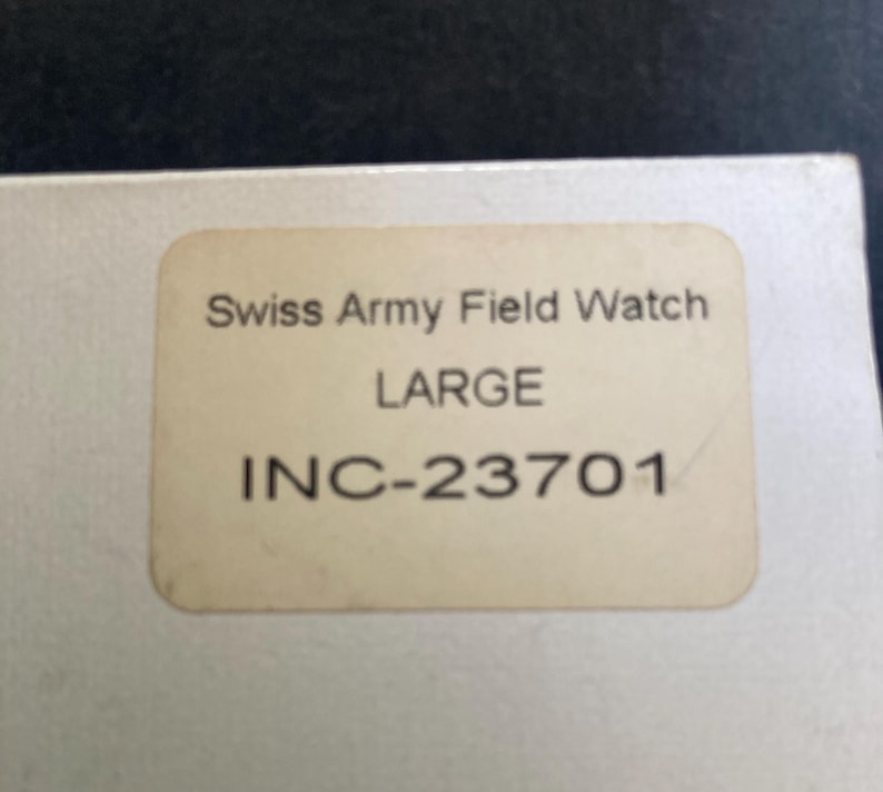 May include: A close-up of a beige label with the text "Swiss Army Field Watch LARGE INC-23701" in black. The label is slightly worn and has rounded corners. The background is a light color.