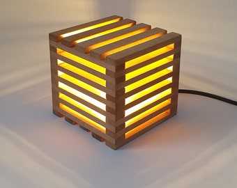 Puede incluir: Una lámpara de madera con forma de cubo y una luz en su interior. La lámpara tiene un brillo cálido y amarillo y está hecha de madera natural.