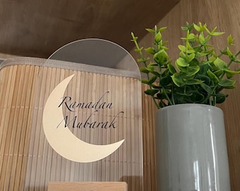Op de afbeelding: Een helder acryl Ramadan Mubarak-bord met een maansikkelontwerp in beige, gemonteerd op een kleine houten basis. Het bord wordt weergegeven voor een geweven mand en een kleine potplant.