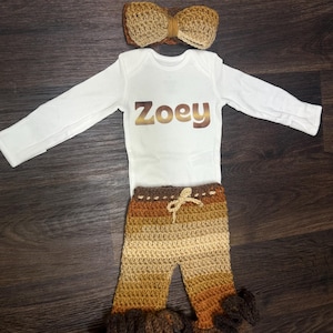 May include: A baby outfit set featuring a white long-sleeve onesie with the name "Zoey" in gold lettering, paired with crochet pants in shades of brown and a matching bow headband. The pants have a drawstring waist and ruffled cuffs.