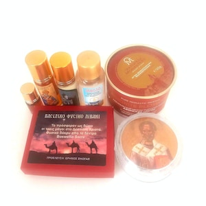 May include: An assortment of religious items, including small bottles with gold caps, a red box with Greek text and camel silhouettes, and a round container with an image of a religious figure. The items are arranged on a white surface.