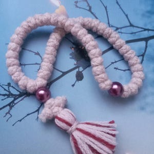 Könnte beinhalten: Ein handgefertigter, cremefarbener und roter Quastenanhänger mit einer passenden Perlenschnur. Der Quastenanhänger ist aus dickem Garn gefertigt und weist ein dekoratives Design auf. Der Hintergrund ist ein blau getöntes Bild mit einer Kerze und Ästen.