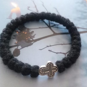 Op de afbeelding: Een zwarte armband van geknoopt koord met een zilverkleurige metalen kruishanger. De armband is opgebouwd uit een reeks uniforme, getextureerde zwarte vezelknopen die in een cirkelvormige lus zijn gerangschikt met een kruisje.