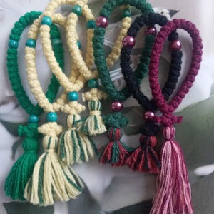 May include: A collection of five knotted cord bracelets featuring various color combinations including green, cream, dark green, black, and burgundy. Each bracelet is accented with round beads and finished with a decorative yarn tassel at the base.