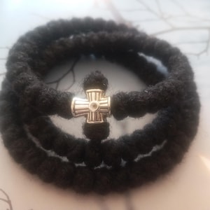 May include: A black prayer rope with a silver cross. The rope is made of thick, woven material and is looped in a circular shape. The cross is centered and has a detailed design.