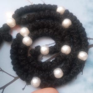 May include: A handmade black rosary with white pearl-like beads. The rosary is made of a thick, textured material and features a cross at the end. The beads are evenly spaced along the rosary's length. The rosary is a religious item.