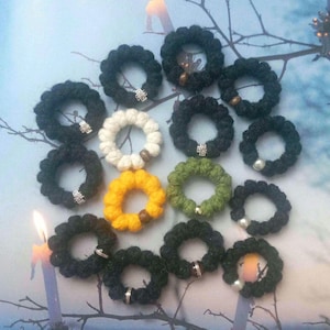 May include: A collection of circular finger rosaries made from knotted yarn in black, white, yellow, and olive green. Each ring features a small decorative accent, including silver-toned cross charms, wooden beads, and white pearl-like beads.