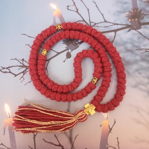 May include: A red prayer rope with gold detailing and a tassel. The knotted rope has gold-coloured spacers. A gold cross pendant is attached to the tassel. The background features lit candles and bare tree branches.