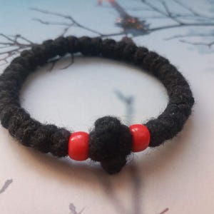 May include: A black prayer rope bracelet with two red beads. The bracelet is made of a thick, woven material and features a cross-shaped knot. The beads are a bright, solid red color.