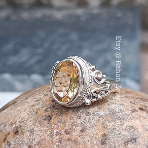 Pode incluir: Um anel de prata com uma pedra preciosa oval, castanho-clara. O anel tem um design ornamentado de filigrana em torno da pedra preciosa e na banda. O anel é exibido em uma superfície de pedra áspera e castanha. O texto diz "Etsy @ Rehan Jewel Hub".