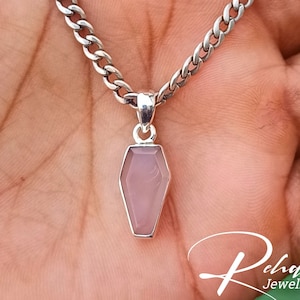 May include: A silver chain necklace with a pink coffin-shaped pendant. The pendant is made of a clear stone with a pink hue.