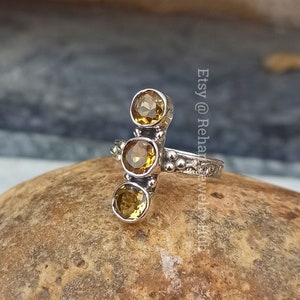 Pode incluir: Anel de prata com três pedras preciosas amarelas dispostas em linha vertical. O anel tem uma banda texturizada.