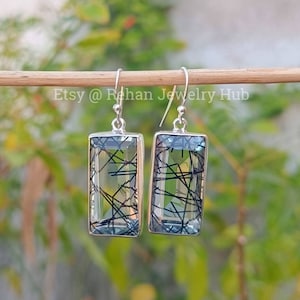 Pode incluir: Brincos de prata com pedras retangulares de quartzo azul e preto. Os brincos estão pendurados em um bastão de madeira. O texto 'Etsy @ Rehan Jewelry Hub' está escrito no bastão.