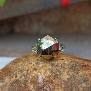 Pode incluir: Um anel de prata com uma gema hexagonal de pirita. O anel está colocado em uma rocha marrom.
