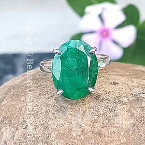 Peut inclure: Une bague en argent avec une grande émeraude de forme ovale. La bague est posée sur une pierre avec une fleur floue en arrière-plan.