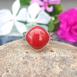 May include: A silver ring with a square red coral gemstone. The ring is set on a gray rock with pink and white flowers in the background.