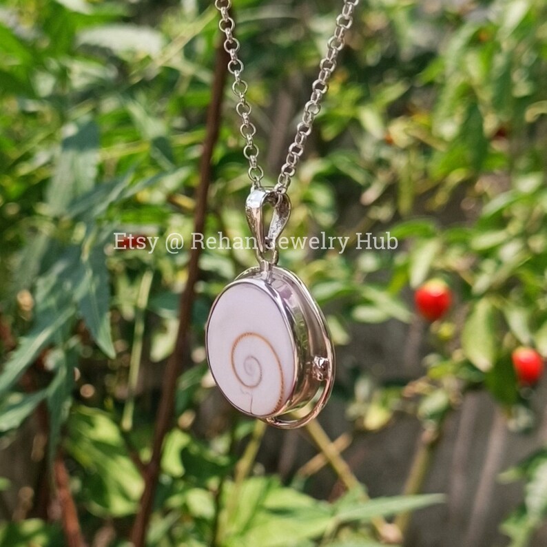 May include: A silver chain necklace with a pendant featuring a white seashell with a brown spiral. The pendant is oval shaped and has a silver frame.