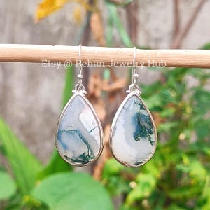 Puede incluir: Un par de pendientes en forma de lágrima con un marco de metal plateado. Los pendientes presentan una piedra con un diseño blanco y verde. Cuelgan de una vara de madera.