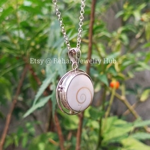 May include: A silver chain necklace with a round pendant featuring a white seashell with a brown spiral pattern. The pendant is set in a silver frame and rotates.