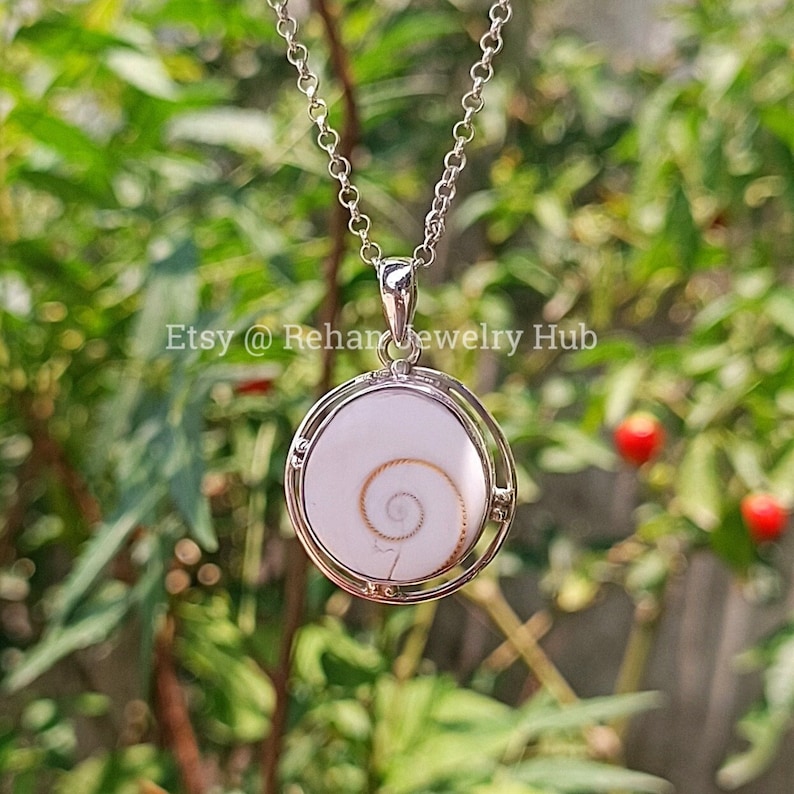 May include: A silver chain necklace with a round pendant featuring a white seashell with a golden spiral inside. The pendant is surrounded by a silver frame.