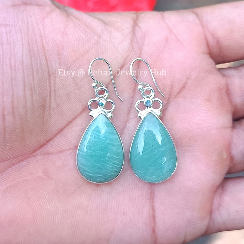May include: A pair of silver dangle earrings with green teardrop-shaped stones and a silver flower design. Etsy @ Rehan Jewelry Hub