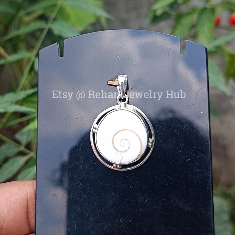 May include: Silver pendant featuring a white seashell with a spiral pattern. The pendant is on a black display stand. Etsy @RehanJewelryHub is printed on the stand.