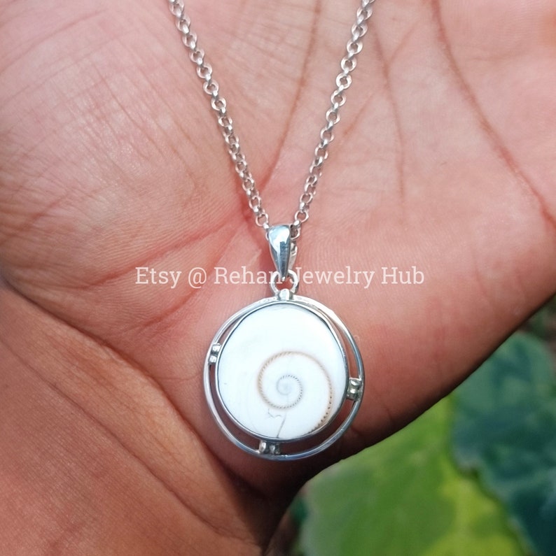 May include: A silver chain necklace with a round pendant featuring a white seashell with a brown spiral inside. The pendant is set in a silver frame with four small silver accents.