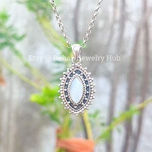 May include: A silver pendant necklace with an oval, pale stone set in an elaborate frame. The frame has a beaded edge and a dark, oxidised finish. The chain is a delicate, linked design. The Etsy shop name "Etsy @ RehanJewelry Hub" is visible.