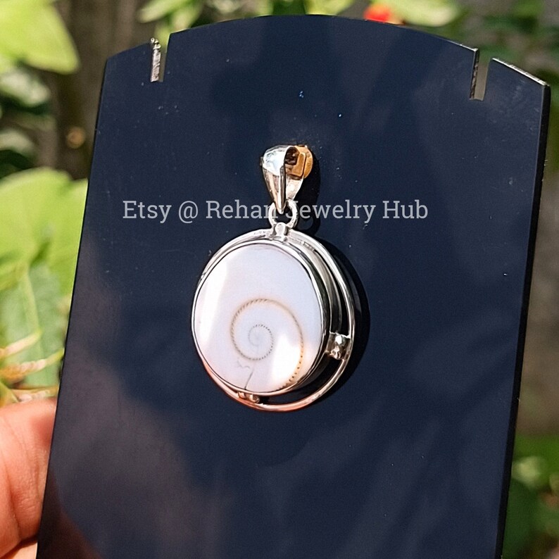 May include: A silver pendant with a white seashell featuring a spiral pattern. The pendant is on a black display stand. Etsy @ Rehan Jewelry Hub.