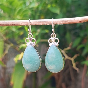 May include: A pair of silver teardrop earrings with light blue green gemstone drops. The earrings are hanging from a wooden surface.