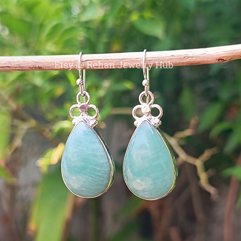 May include: A pair of silver teardrop earrings with light blue green gemstone drops. The earrings are hanging from a wooden surface.
