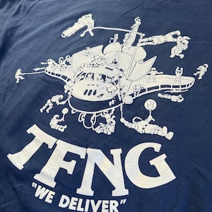 May include: A navy blue t-shirt with a white graphic of a spaceship and cartoon astronauts. The text "TFNG" and "WE DELIVER" is printed in white on the shirt.