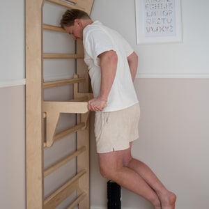 May include: A wooden stall bar unit with horizontal rungs is mounted on a wall. A person is using the stall bars for exercise. A black foam roller is on the floor. A framed alphabet print hangs on the wall.