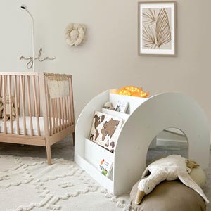 May include: A white wooden bookshelf with a curved archway and shelves for books and toys. The bookshelf is decorated with a world map and a stuffed animal. The bookshelf is in a nursery with a crib and a rug.