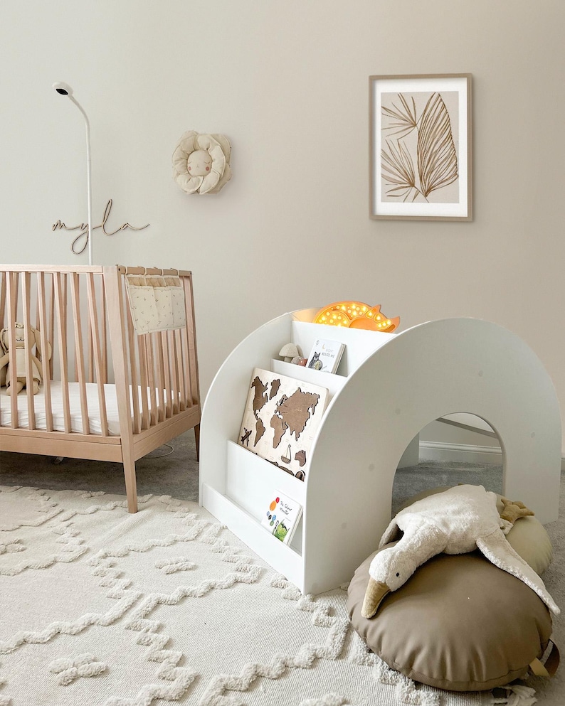 May include: A white wooden bookshelf with a curved archway and shelves for books and toys. The bookshelf is decorated with a world map and a stuffed animal. The bookshelf is in a nursery with a crib and a rug.