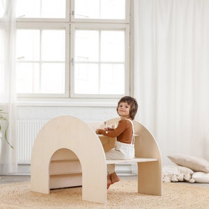 May include: A light wood children's play table and chair set with a curved, arched design. The table has a shelf underneath and the chair has a small back. A child is sitting at the table.