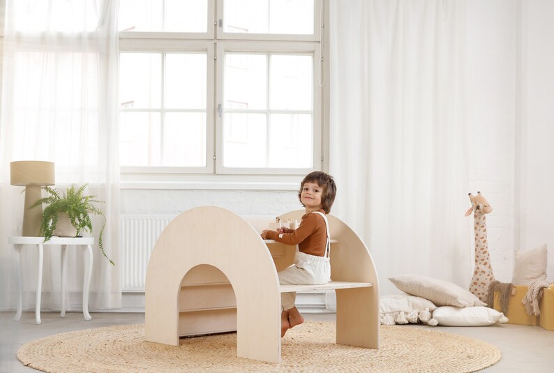 May include: A light wood children's play table and chair set with a curved, arched design. The table has a shelf underneath and the chair has a small back. A child is sitting at the table.