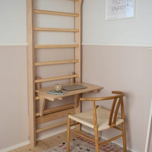 Puede incluir: Un espaldera de madera montada en la pared con un escritorio plegable, un portátil y una taza pequeña. Una silla tejida está sobre una alfombra estampada. Una impresión enmarcada del alfabeto cuelga de la pared.