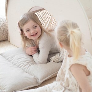 May include: Two young girls playing in a white wooden playhouse with a beige cushion. The girl in the front is wearing a beige sweater and the girl in the back is wearing a white dress with a floral pattern.