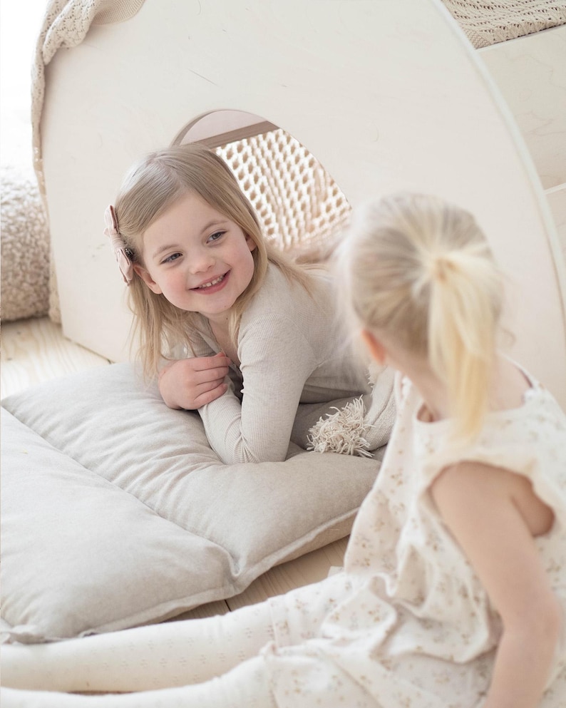 May include: Two young girls playing in a white wooden playhouse with a beige cushion. The girl in the front is wearing a beige sweater and the girl in the back is wearing a white dress with a floral pattern.