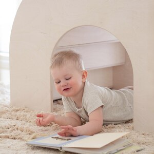 May include: A young child is lying on a beige rug, looking at a book with a blue cover. The child is wearing a light grey shirt and is in a white wooden playhouse.