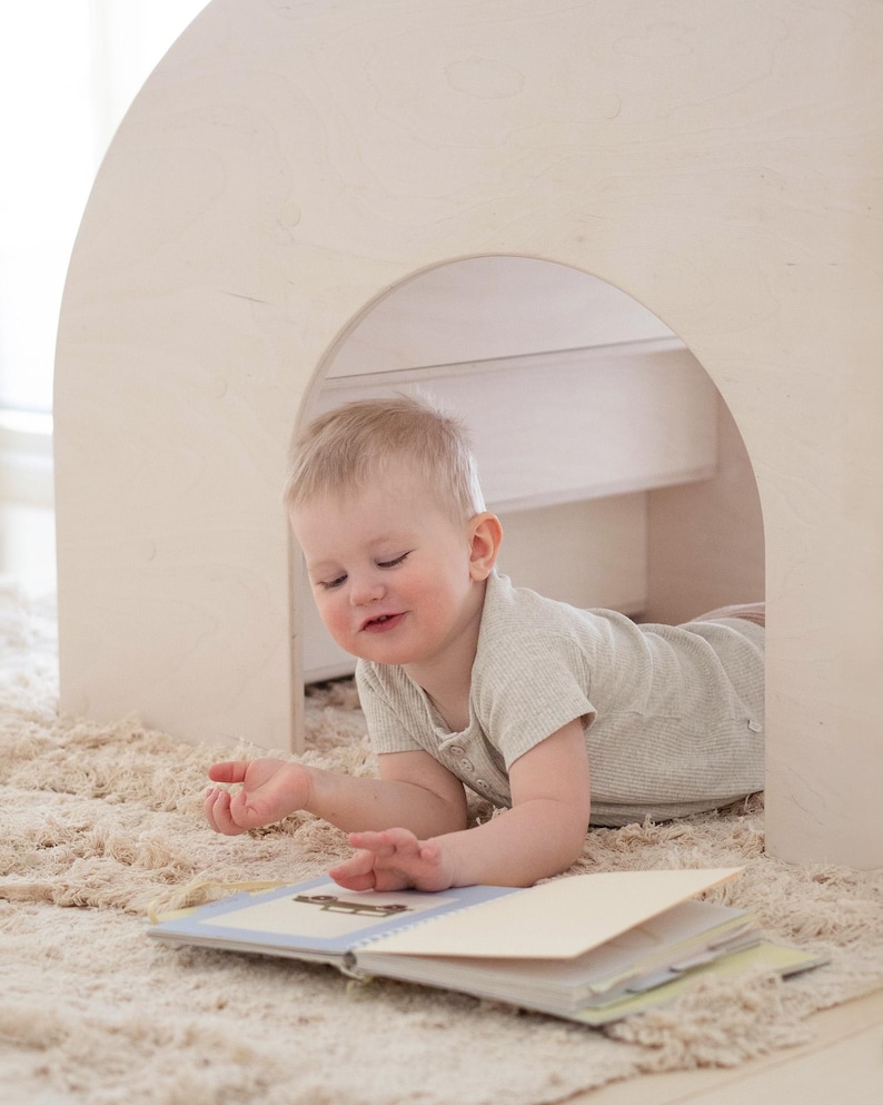 May include: A young child is lying on a beige rug, looking at a book with a blue cover. The child is wearing a light grey shirt and is in a white wooden playhouse.