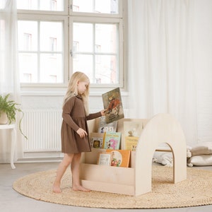 May include: A light wood bookshelf with a rounded top and shelves for books. A young girl in a brown dress is standing next to the bookshelf and holding a book. The bookshelf is on a light brown rug in a room with white walls and a window.