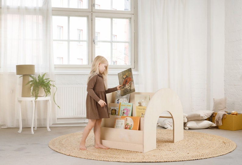 May include: A light wood bookshelf with a rounded top and shelves for books. A young girl in a brown dress is standing next to the bookshelf and holding a book. The bookshelf is on a light brown rug in a room with white walls and a window.