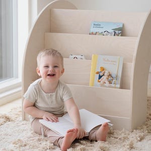 May include: A young child sits on a beige rug in front of a white wooden bookshelf with three shelves. The bookshelf is curved and has a rounded top. The child is holding a white book and smiling. There are three books on the shelves, one with the title "Seilim ja villisimet" and another with the title "Kultaa puuma!"