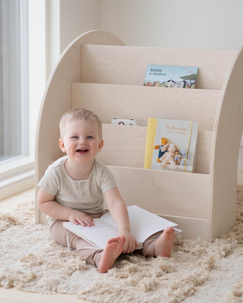 May include: A young child sits on a beige rug in front of a white wooden bookshelf with three shelves. The bookshelf is curved and has a rounded top. The child is holding a white book and smiling. There are three books on the shelves, one with the title "Seilim ja villisimet" and another with the title "Kultaa puuma!"