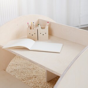 May include: A wooden children's table with a rounded edge and a shelf. The table has two small wooden containers with animal faces on them, holding colored pencils. A blank notebook is on the table.
