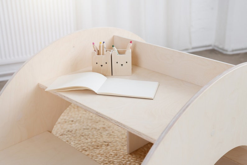 May include: A wooden children's table with a rounded edge and a shelf. The table has two small wooden containers with animal faces on them, holding colored pencils. A blank notebook is on the table.
