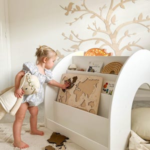 May include: A white wooden bookshelf with a built-in playhouse, featuring a world map puzzle and a child playing with the puzzle pieces. The bookshelf has three shelves and a curved top. The playhouse has a small opening and a soft cushion inside.
